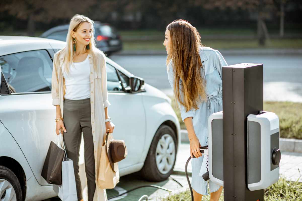 Two smartly dressed women holding shopping bags. One is plugging her electric vehicle into a charger.